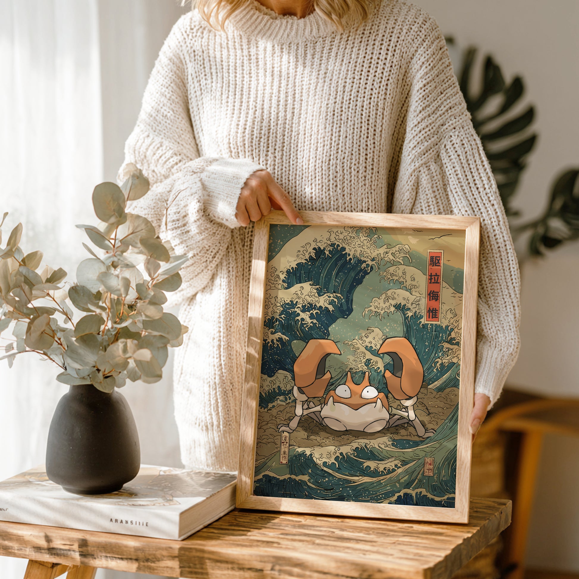 Person holding a framed artwork of a stylized Krabby Pokemon on a wave, with a vase and plant in the background.