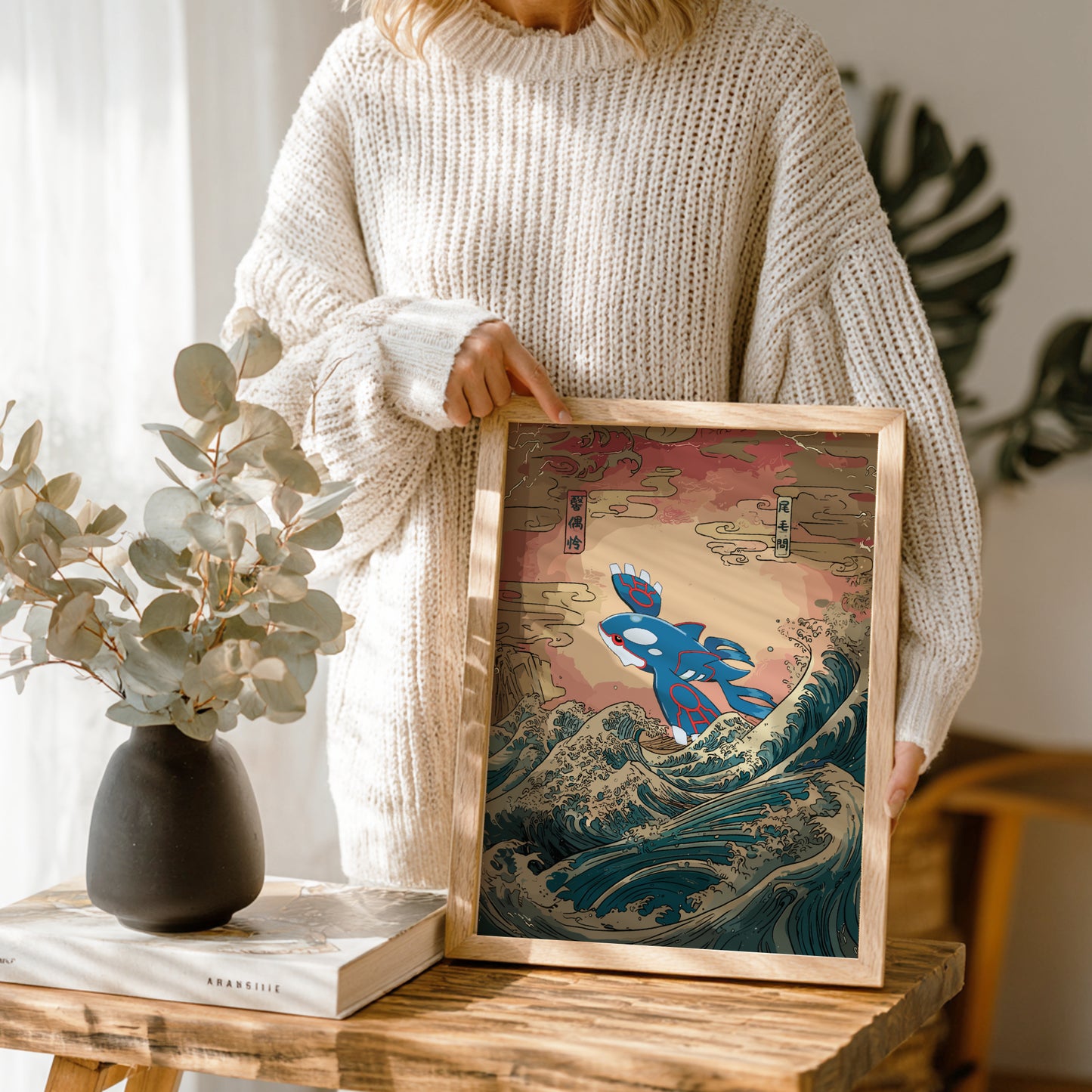 Person holding a framed artwork of Kyogre Pokemon on a wave, standing next to a vase with greenery.