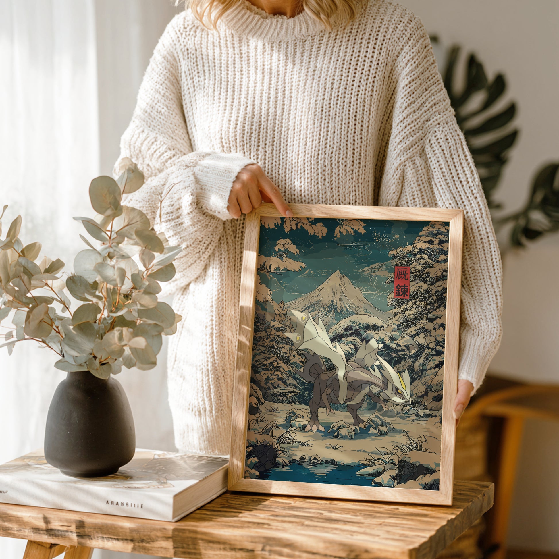 Person holding a framed artwork of a mountain landscape with Kyurem Pokemon, standing next to a vase and book on a wooden table.