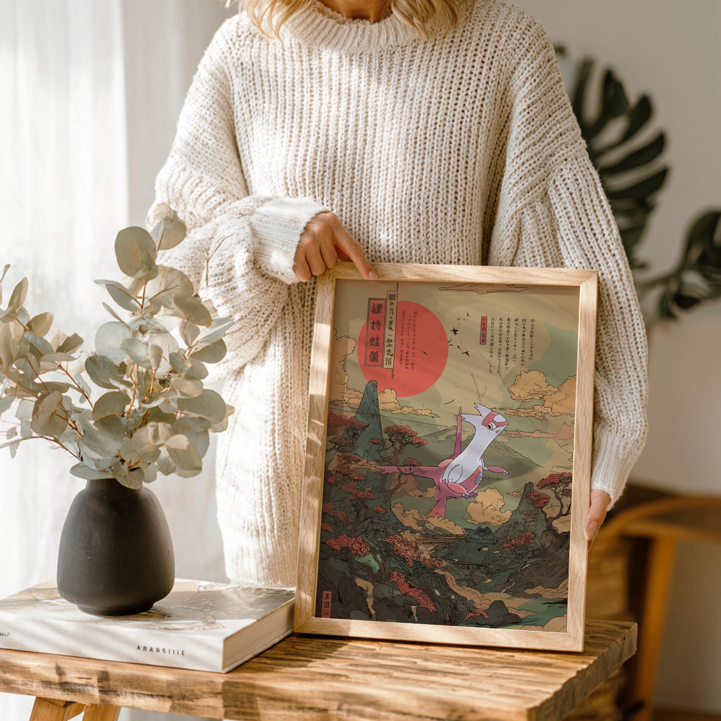 Person holding a framed artwork of Latias Pokemon in a scenic setting, with a vase and book on a wooden table.
