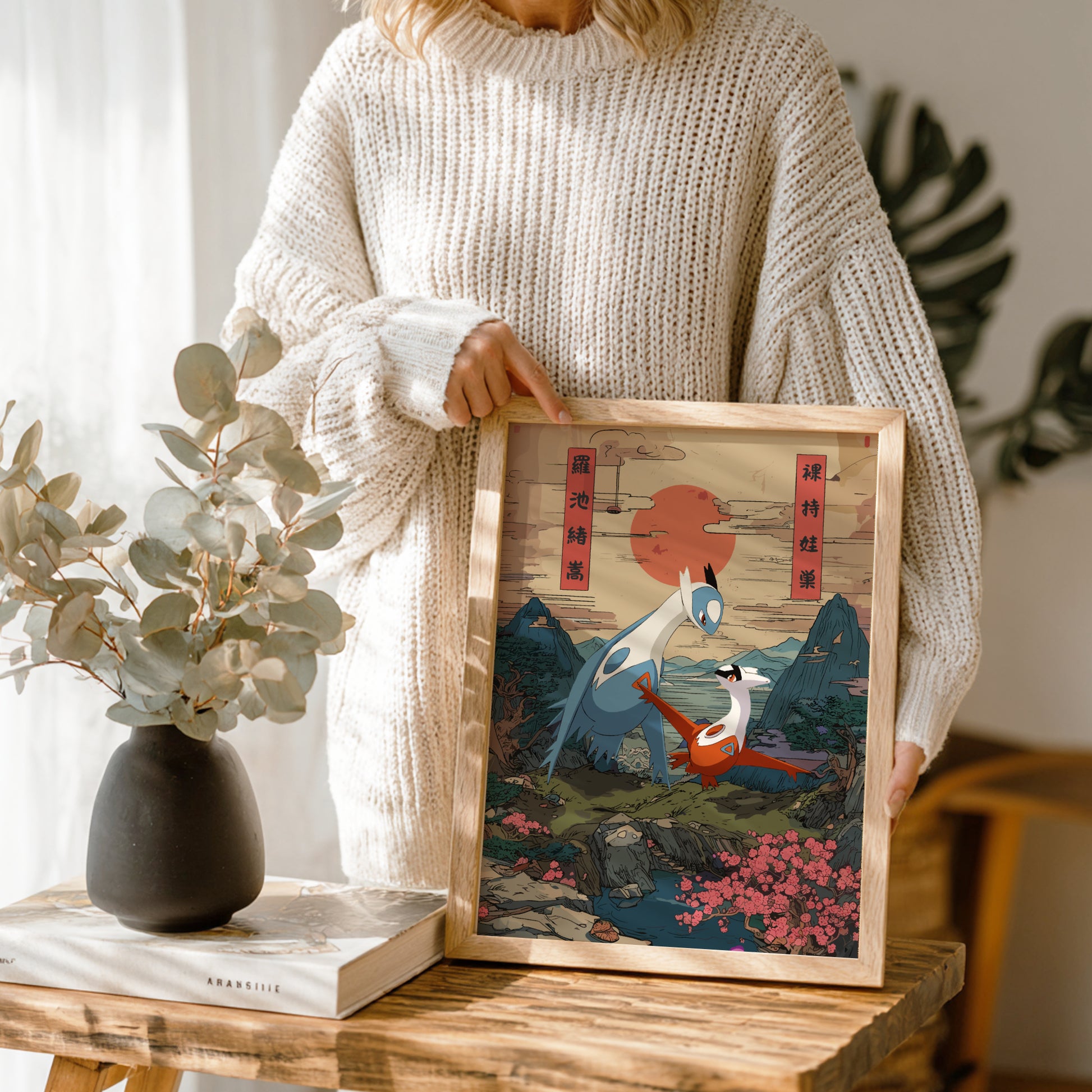 Person holding a framed artwork with a mountain and Latias and Latios Pokemon design, standing next to a vase and books on a wooden table.