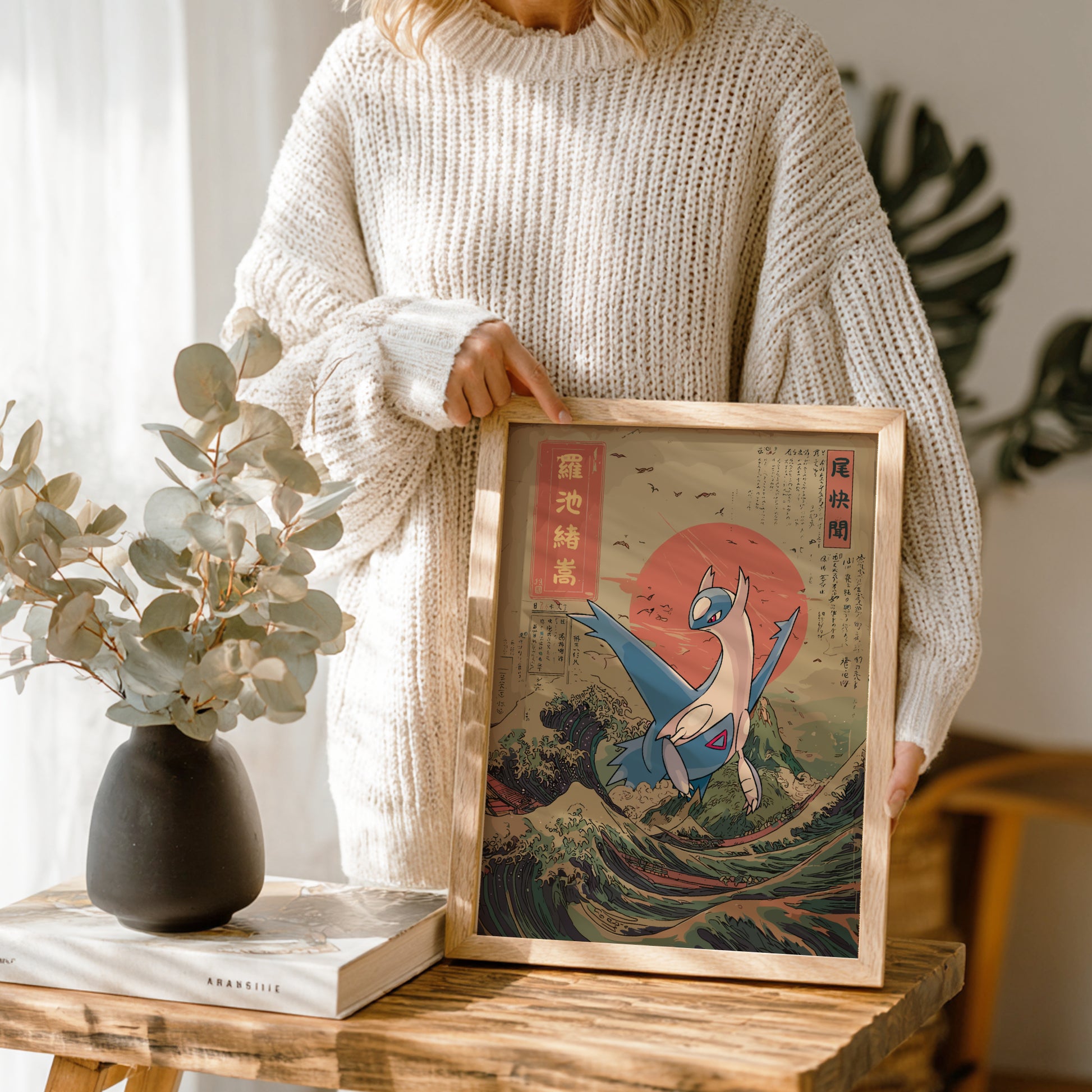 Person holding a framed artwork of Latios Pokemon on a wave with a vase and book in the background