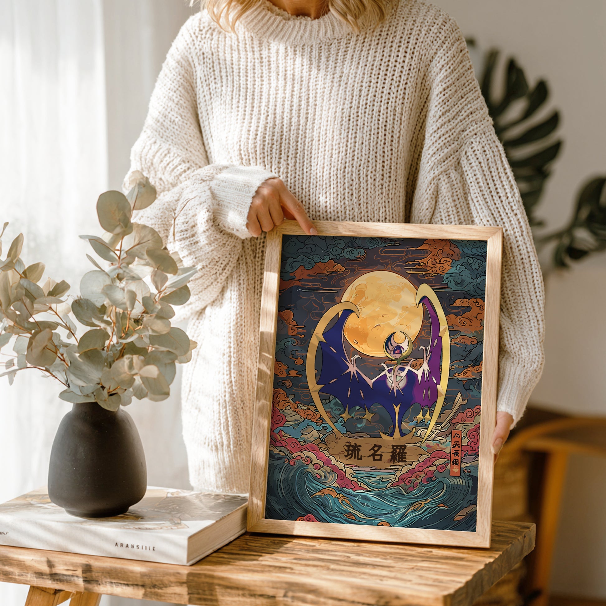 Person holding a framed artwork of Lunala Pokemon with a stylized moon and wave design, standing in a home setting.