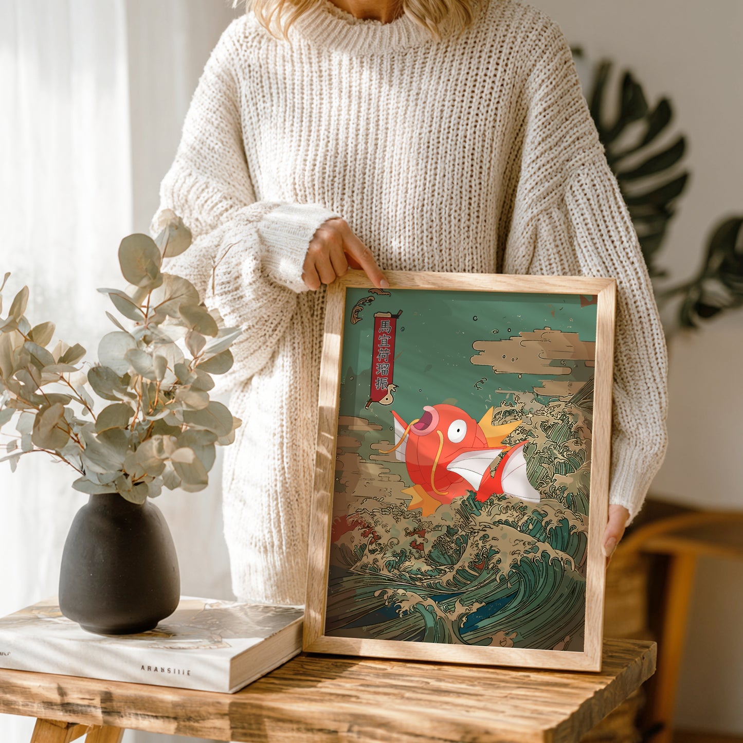 Person holding a framed artwork of Magikarp Pokemon on a wooden table with a vase and book in the background.