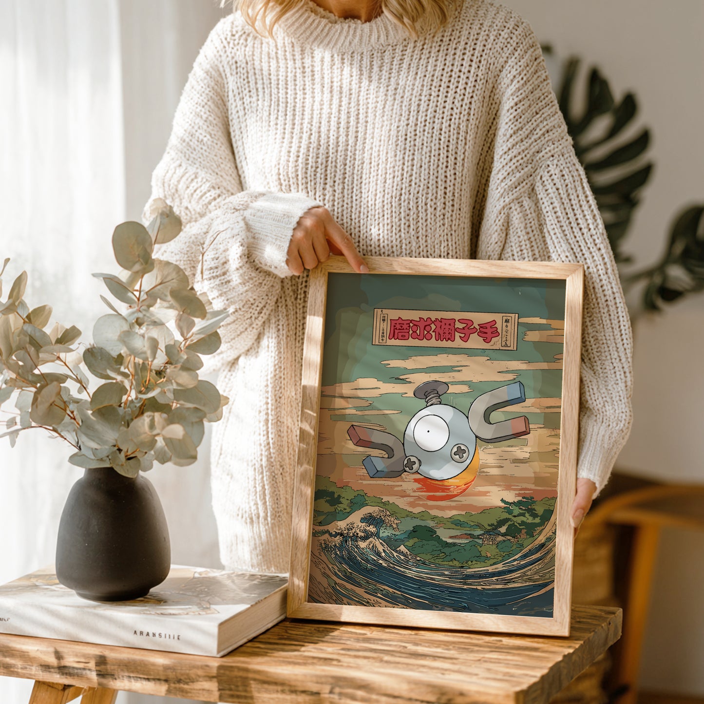 Person holding a framed artwork with a colorful design of Magnemite Pokemon on a wooden table.
