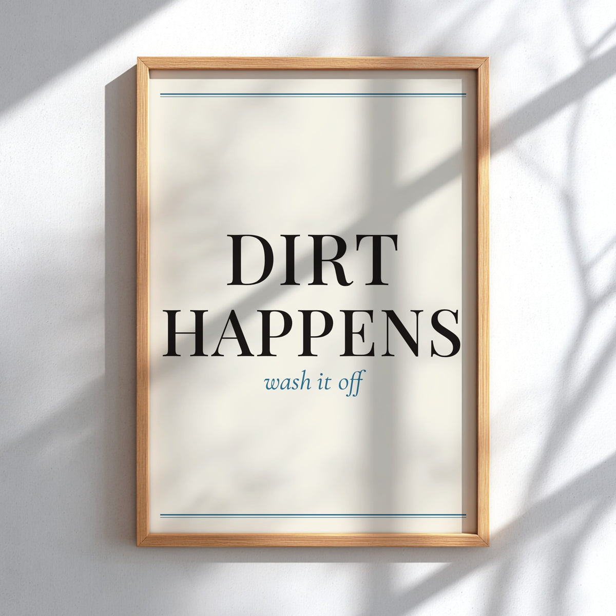 A framed typographic wall art print hanging on a white wall, with soft shadow patterns cast across it. The print reads “DIRT HAPPENS” in large black serif letters and “wash it off” in smaller blue script below, with thin blue horizontal lines near the top and bottom.