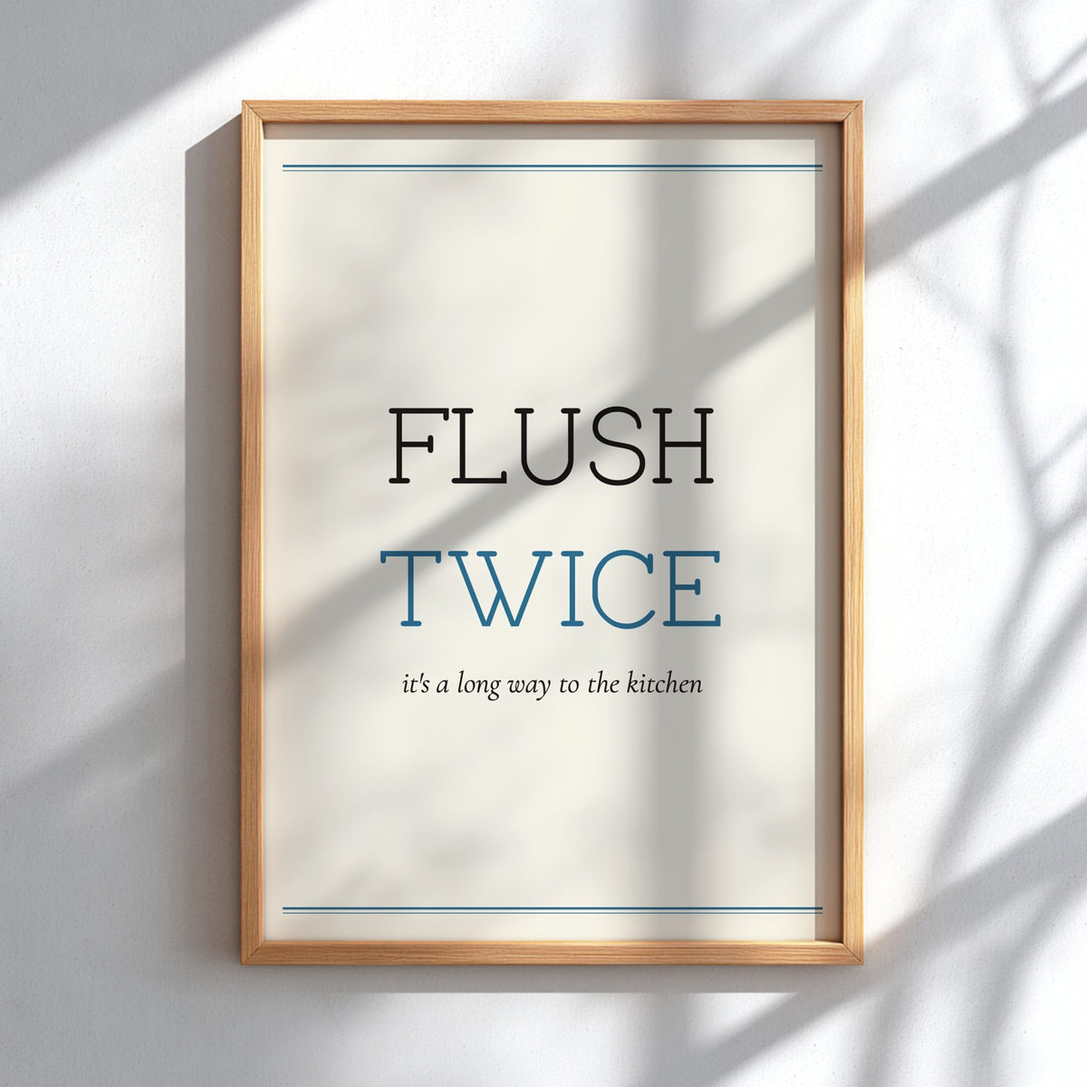 A framed typographic wall art print reading “FLUSH TWICE” with smaller text “it’s a long way to the kitchen,” shown in a light wood frame against a white wall with soft shadow overlays.
