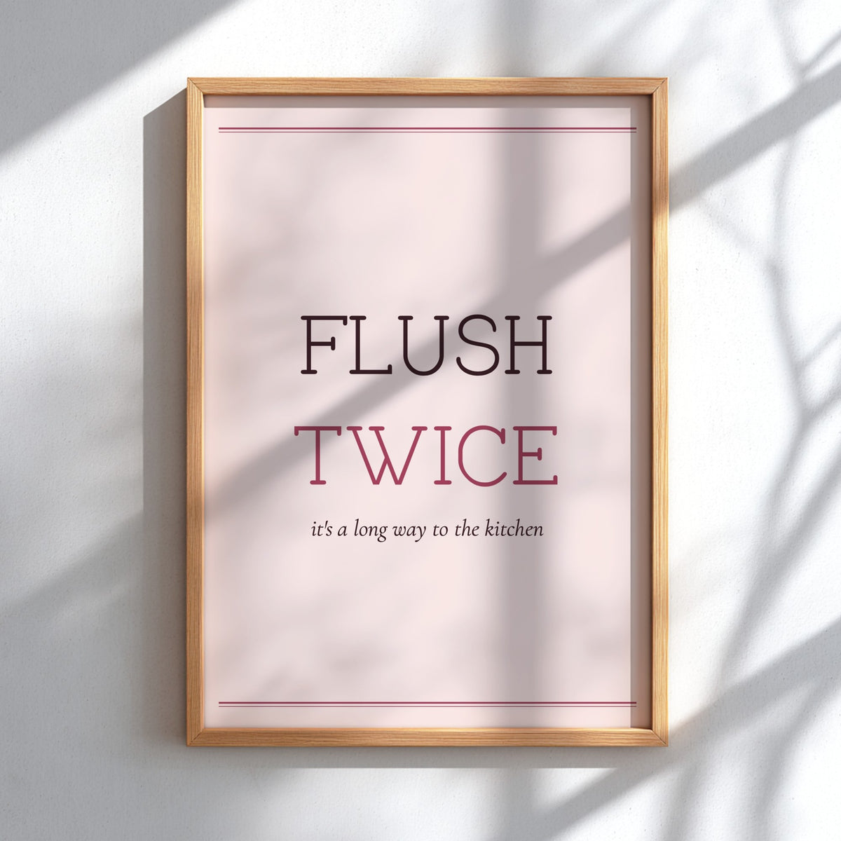 A framed typographic wall art print reading “FLUSH TWICE” with smaller text “it’s a long way to the kitchen,” shown in a light wood frame against a white wall with soft shadow overlays.