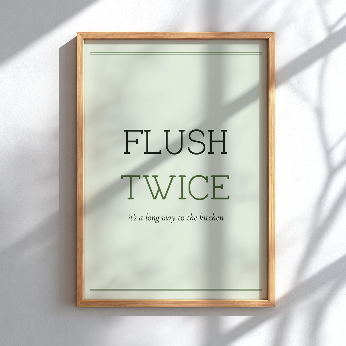 A framed typographic wall art print reading “FLUSH TWICE” with smaller text “it’s a long way to the kitchen,” shown in a light wood frame against a white wall with soft shadow overlays.