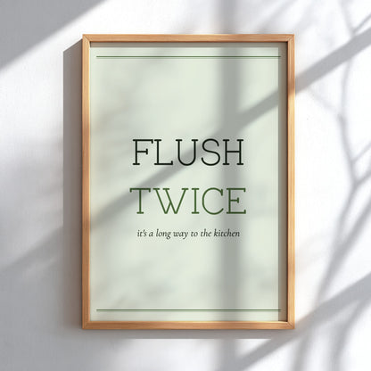A framed typographic wall art print reading “FLUSH TWICE” with smaller text “it’s a long way to the kitchen,” shown in a light wood frame against a white wall with soft shadow overlays.
