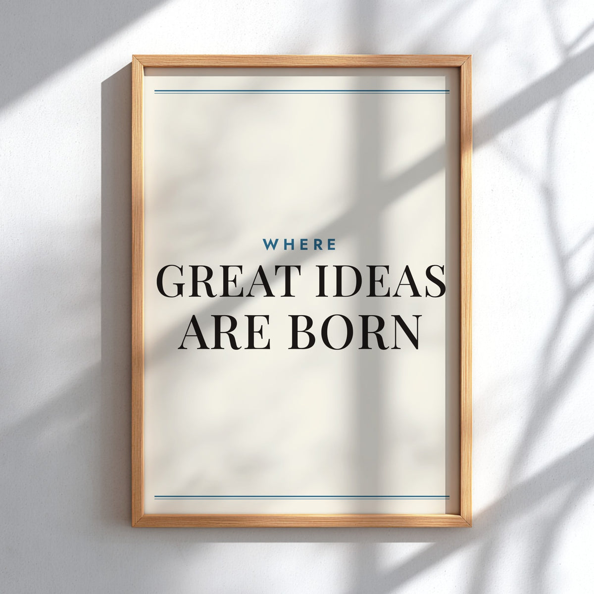 A framed typographic wall art print with the text 'WHERE GREAT IDEAS ARE BORN' in black and blue lettering on an off-white background, shown in a light wood frame against a white wall with soft shadow patterns.