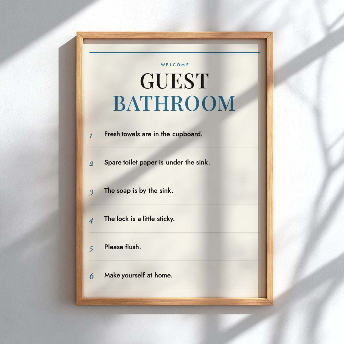 A framed guest bathroom instruction poster hanging on a light gray wall, with soft diagonal shadows cast across it. The poster has a cream background with black and blue text and is enclosed in a light natural wood frame.