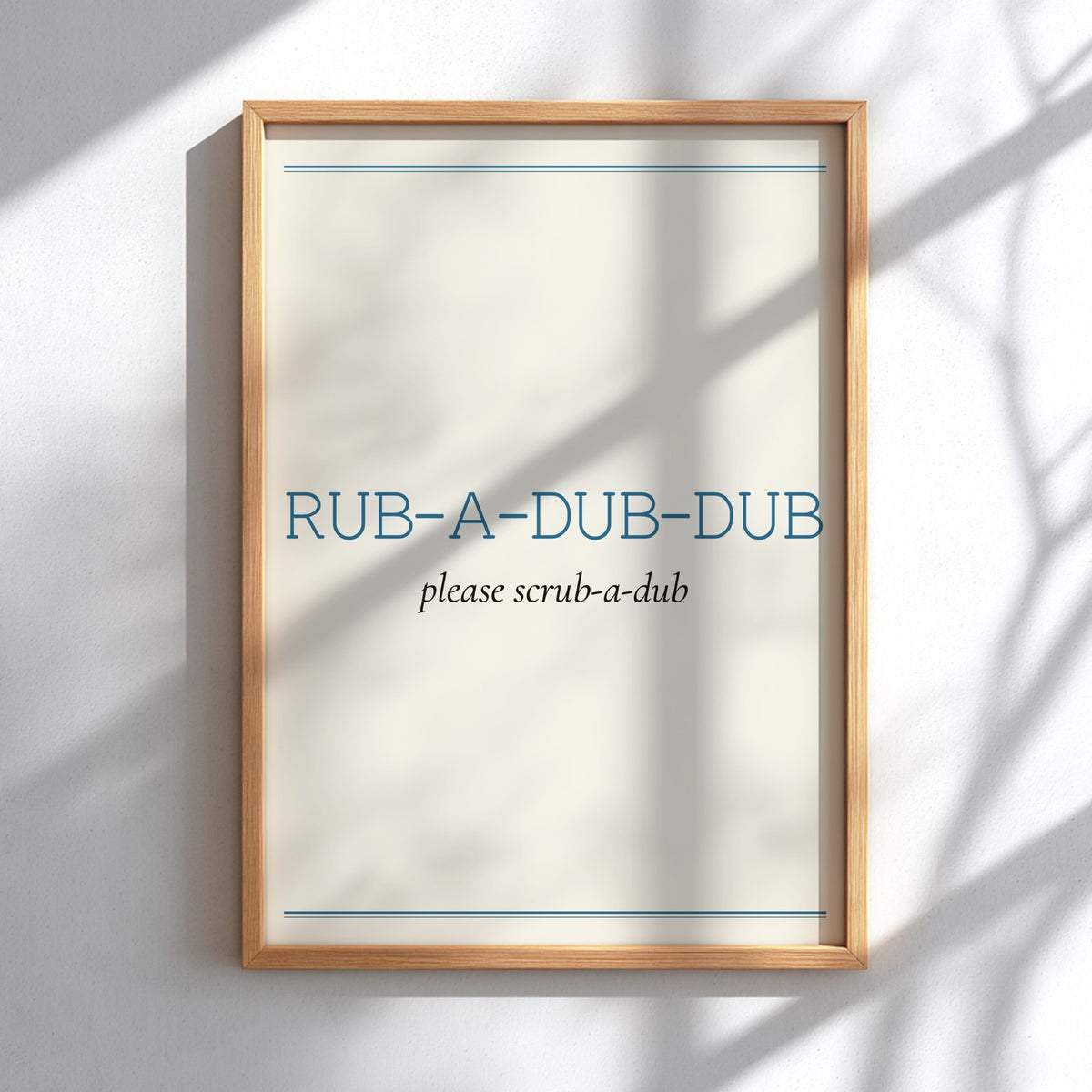 A light wood framed poster with the text 'RUB-A-DUB-DUB' and 'please scrub-a-dub' in blue and black lettering, displayed on a white wall with soft shadow patterns.