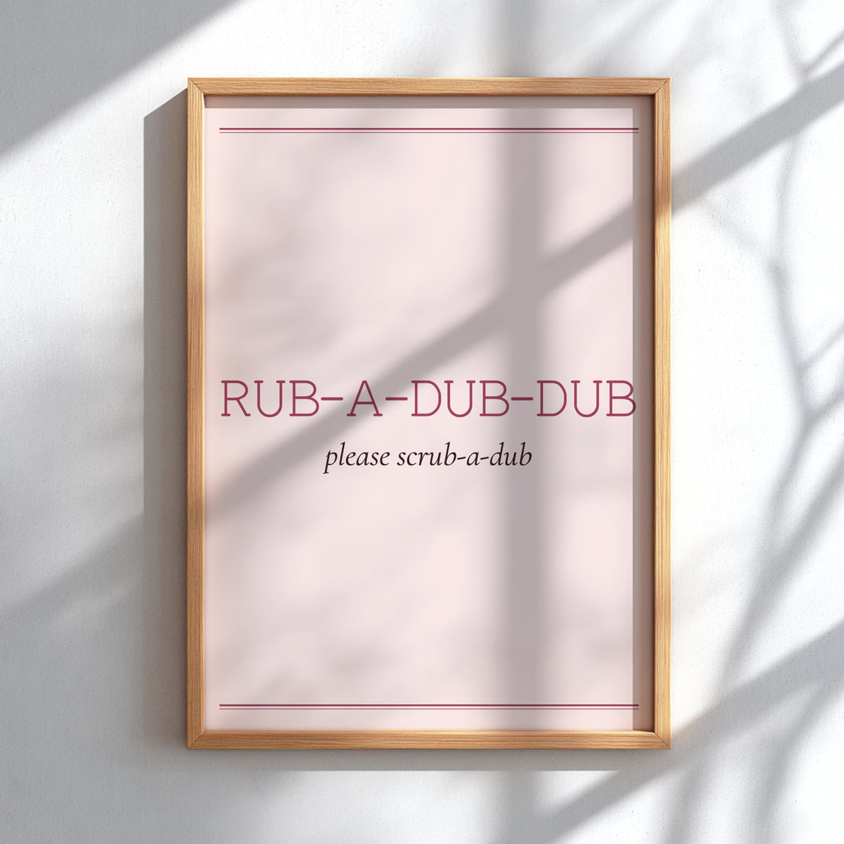 A framed wall art print with a light wood frame, featuring the text 'RUB-A-DUB-DUB' and 'please scrub-a-dub' on a pale pink background, mounted on a white wall with soft shadow patterns.