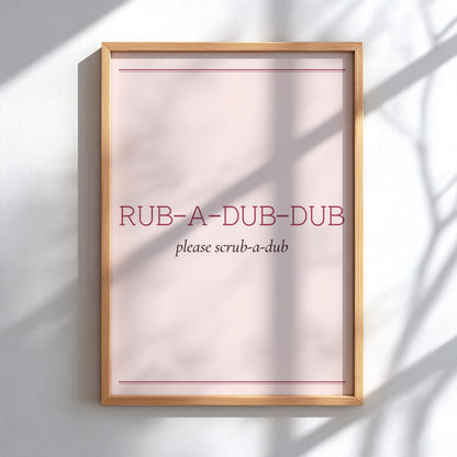 A framed wall art print with a light wood frame, featuring the text 'RUB-A-DUB-DUB' and 'please scrub-a-dub' on a pale pink background, mounted on a white wall with soft shadow patterns.