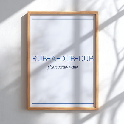 A framed wall sign with a light wood frame and a white background, featuring the blue text 'RUB-A-DUB-DUB' and smaller text 'please scrub-a-dub'. The sign is mounted on a white wall with soft shadow patterns cast across it.
