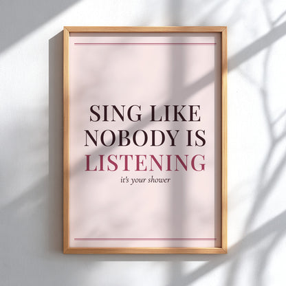 Framed typographic wall art hanging on a white wall, with soft shadow overlays. The print has a pale blush background, dark brown text reading 'SING LIKE NOBODY IS LISTENING', and smaller text 'it's your shower' beneath, bordered by thin pink lines.