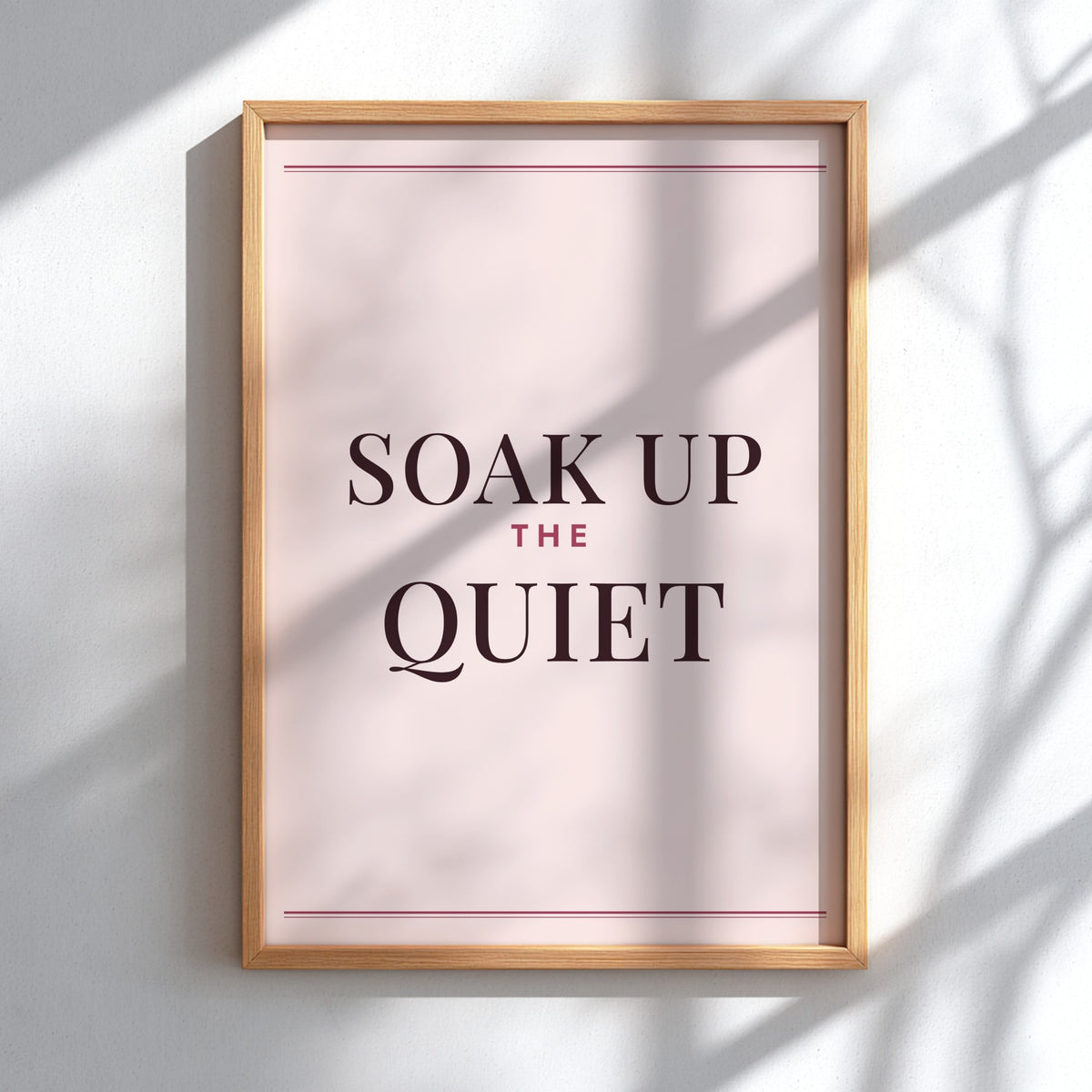 A framed typographic wall art print reading “SOAK UP THE QUIET,” with dark serif text on a pale blush background and a light natural wood frame, mounted on a white wall with soft shadow overlays.