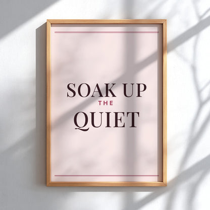 A framed typographic wall art print reading “SOAK UP THE QUIET,” with dark serif text on a pale blush background and a light natural wood frame, mounted on a white wall with soft shadow overlays.