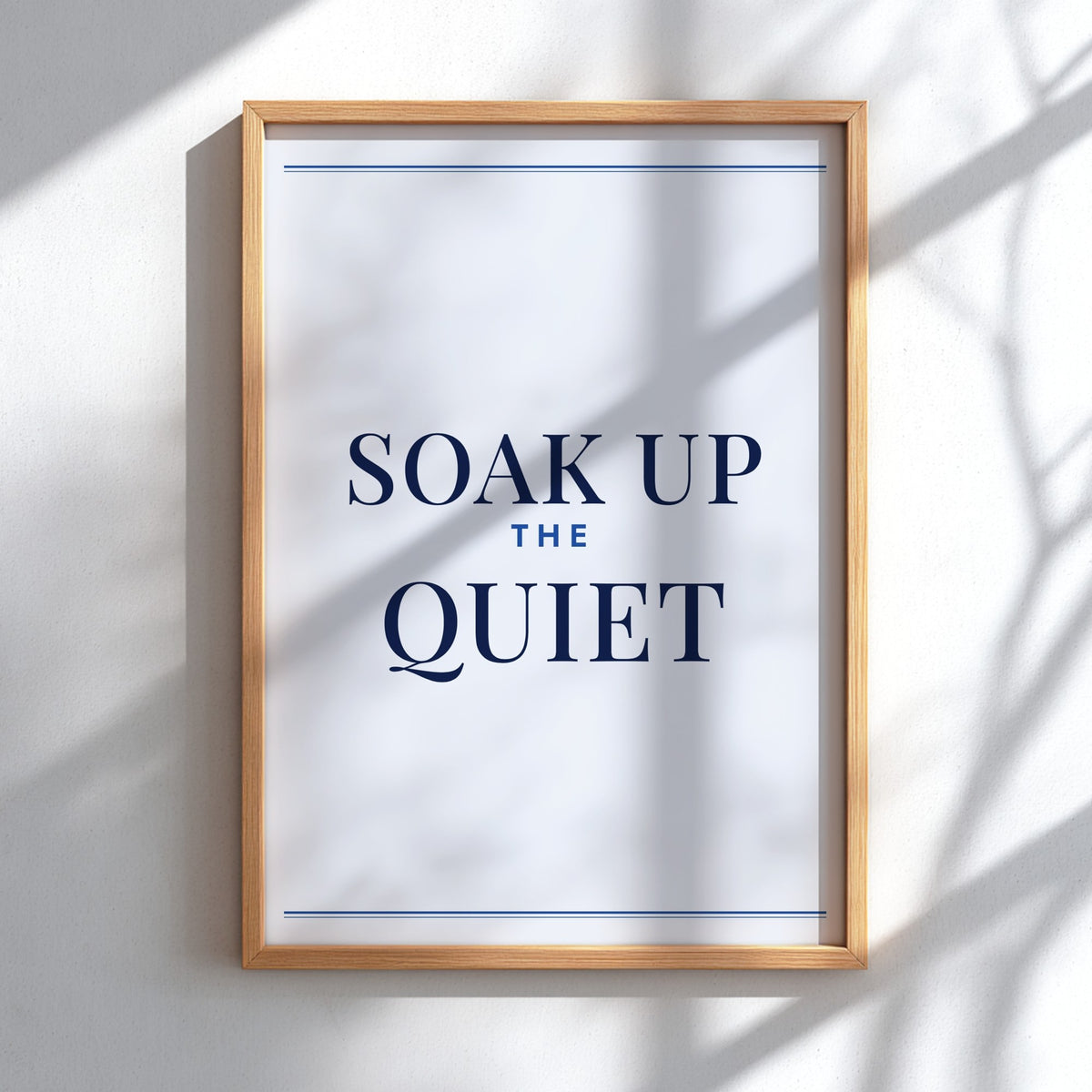 A framed typographic wall art print reading “SOAK UP THE QUIET” in dark navy text on a white background, hung on a light wall with soft shadow overlays.
