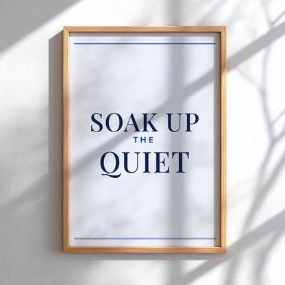 A framed typographic wall art print reading “SOAK UP THE QUIET” in dark navy text on a white background, hung on a light wall with soft shadow overlays.