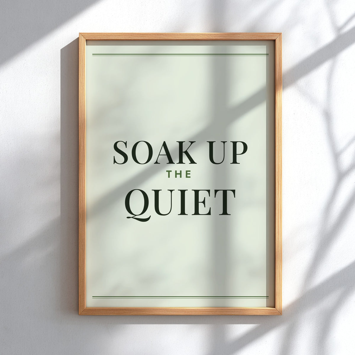 A framed typographic wall art print reading “SOAK UP THE QUIET” in dark green serif text on a pale sage background, mounted on a white wall with soft shadow overlays.