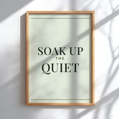 A framed typographic wall art print reading “SOAK UP THE QUIET” in dark green serif text on a pale sage background, mounted on a white wall with soft shadow overlays.
