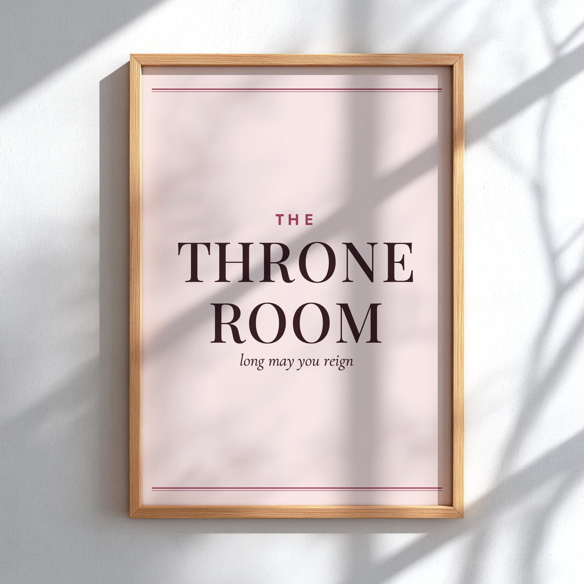 A framed typographic wall art print reading 'THE THRONE ROOM' with smaller text 'long may you reign' on a pale blush background, shown in a light wood frame hanging on a white wall with soft shadow overlays.