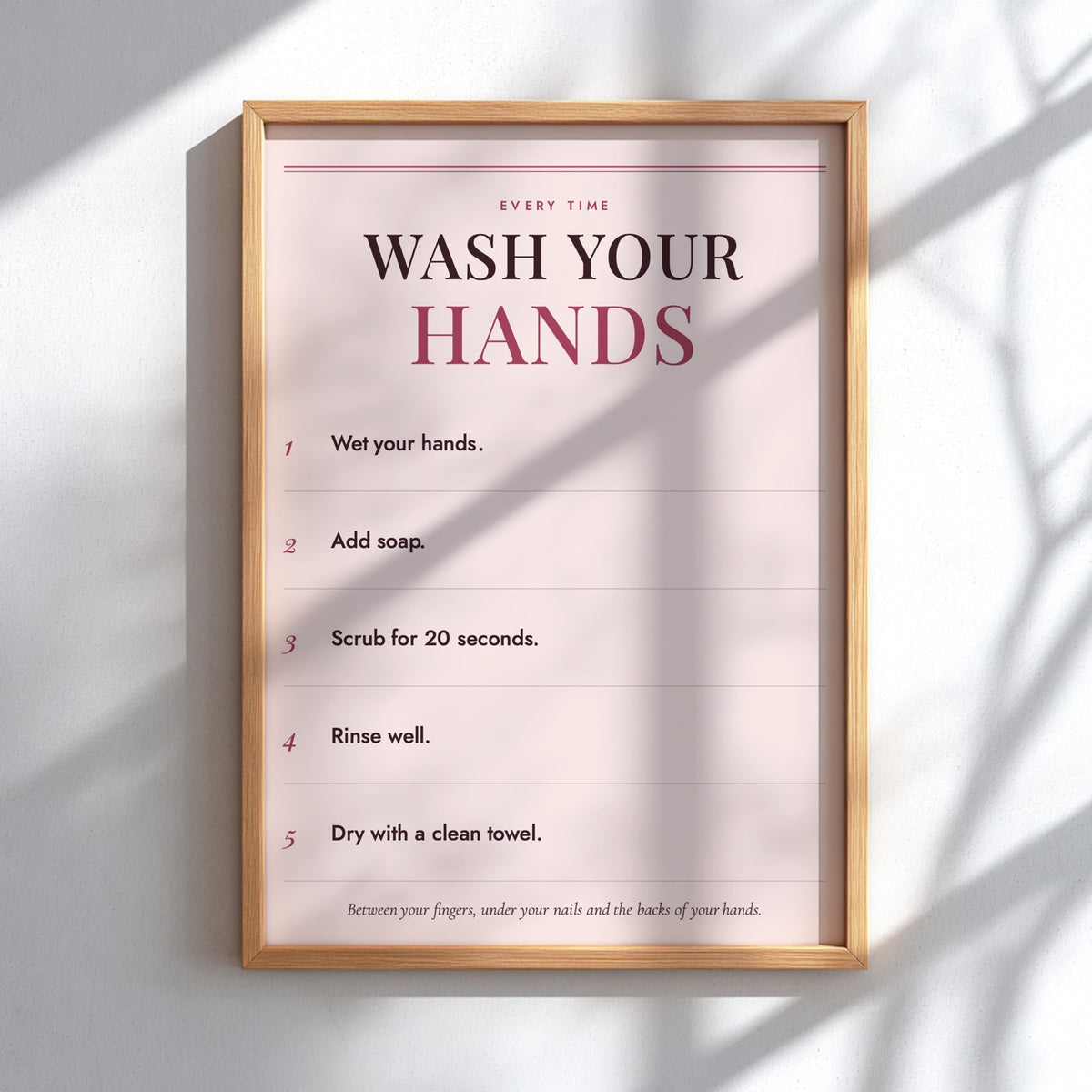 A framed handwashing instruction poster hanging on a white wall, with sunlight casting soft shadows across the print and frame.