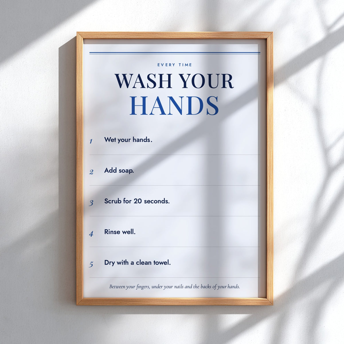 A framed handwashing instruction poster hanging on a white wall, with natural window shadows cast across it. The poster reads 'WASH YOUR HANDS' and lists five steps, enclosed in a light wood frame.