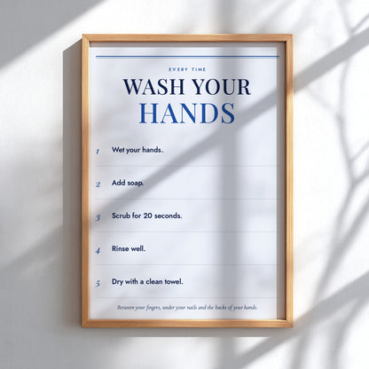 A framed handwashing instruction poster hanging on a white wall, with natural window shadows cast across it. The poster reads 'WASH YOUR HANDS' and lists five steps, enclosed in a light wood frame.