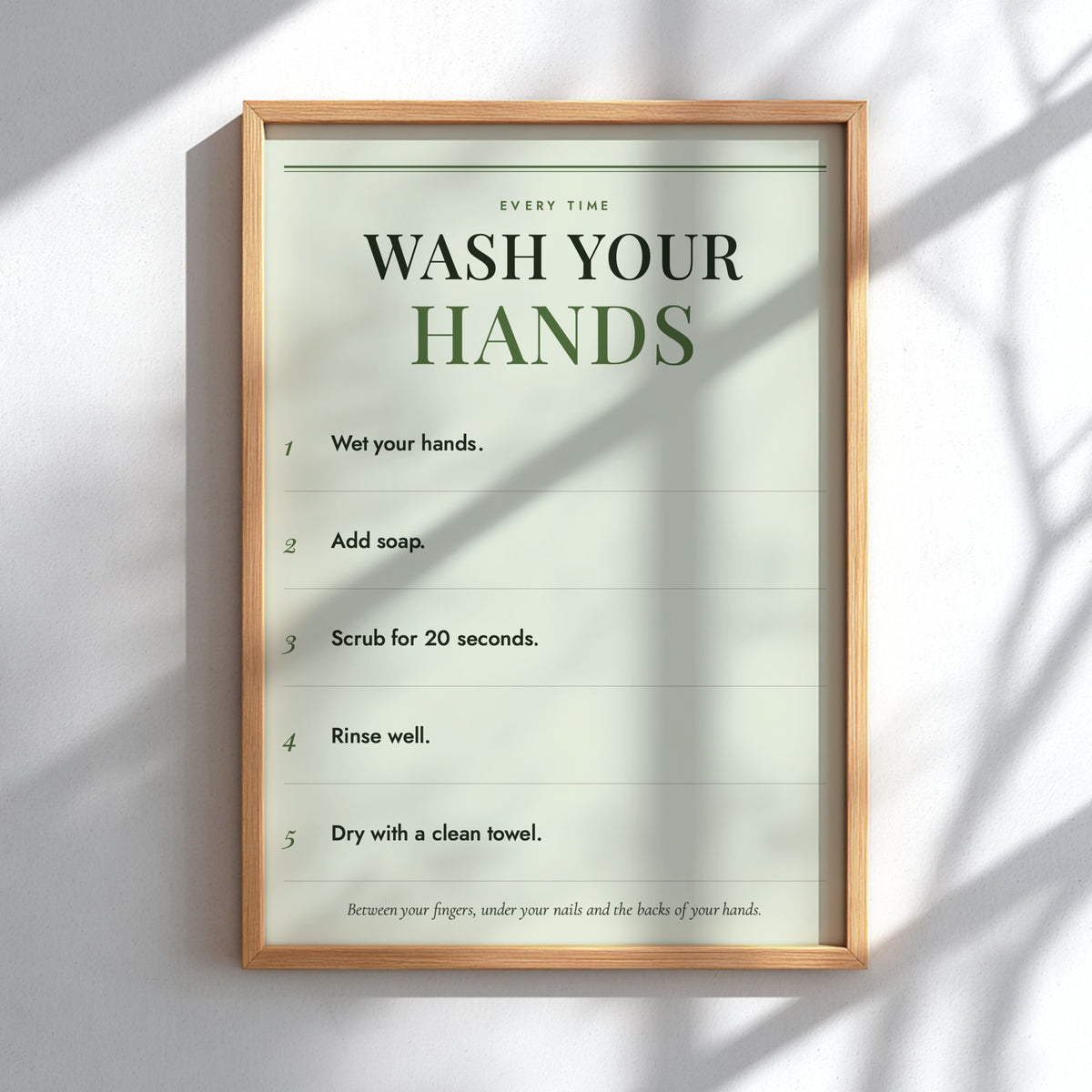 A framed handwashing instruction poster mounted on a white wall, with natural window shadows cast across it. The poster has a light wood frame and a pale green background with dark green and black text reading 'WASH YOUR HANDS' followed by five steps.