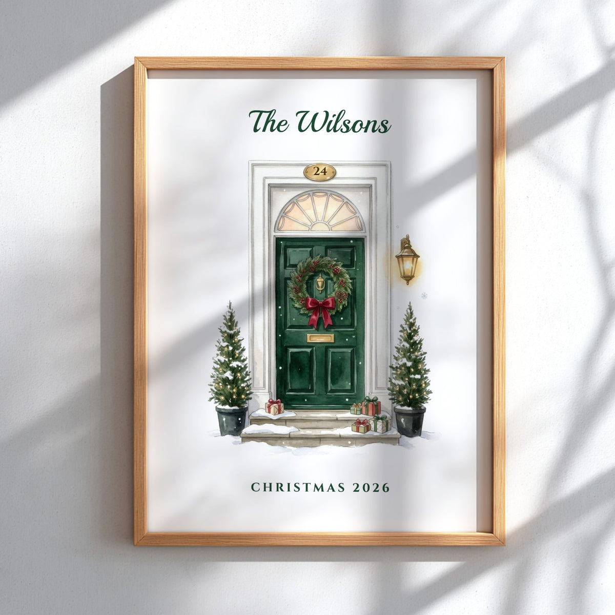 A personalized framed Christmas art print featuring an illustration of a green front door with a wreath, holiday trees, and the text 'The Wilsons' and 'Christmas 2026'. The print is housed in a light wood frame and displayed on a white wall with soft window shadow patterns.