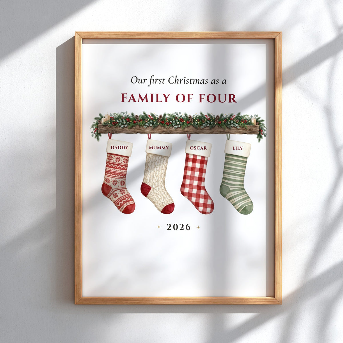 A personalized framed Christmas print on a white wall with soft window shadows. The print features a light wood frame and displays the text 'Our first Christmas as a FAMILY OF FOUR' above four custom-designed Christmas stockings labeled 'DADJOY', 'MUMMY', 'OSCAR', and 'LILY' hanging from a green garland.