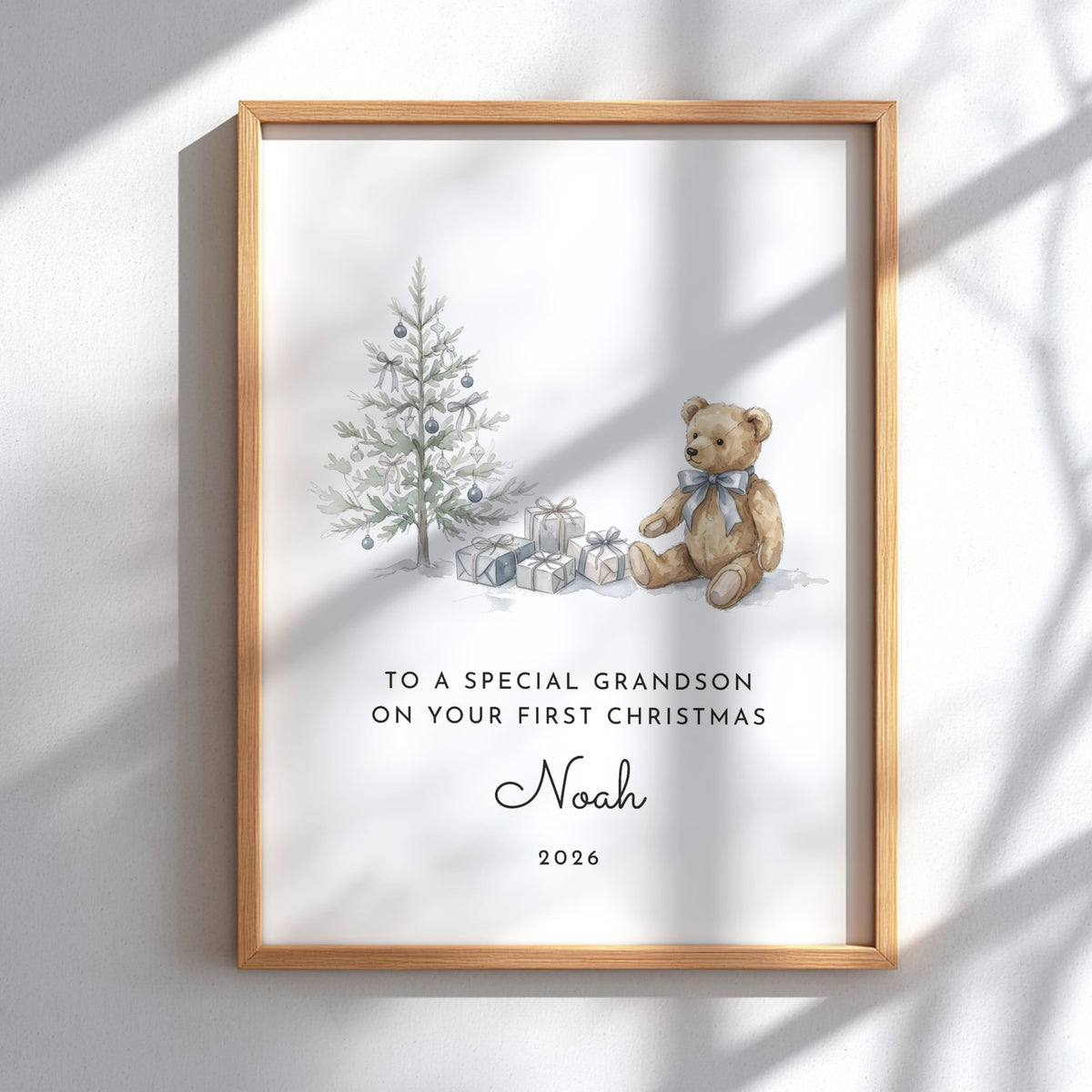 A personalized framed Christmas print featuring a watercolor teddy bear, a decorated tree, and wrapped gifts, with the text 'TO A SPECIAL GRANDSON ON YOUR FIRST CHRISTMAS Noah 2026'. The print is in a light wood frame and displayed against a white wall with soft window shadows.
