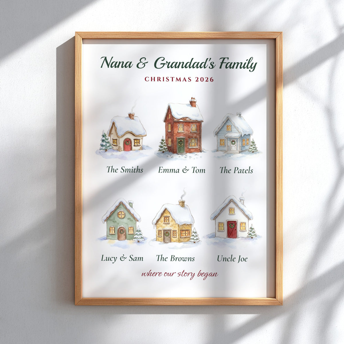 A personalized framed Christmas print featuring six illustrated winter cottages with family names underneath, mounted on a light wood frame and displayed against a white wall with soft shadow overlays.
