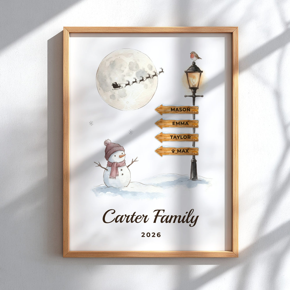 A personalized framed art print featuring a winter night scene with a full moon, Santa Claus, a snowman, and a streetlamp with custom names. The print is in a light wood frame and displayed on a white wall with soft window shadow overlays.