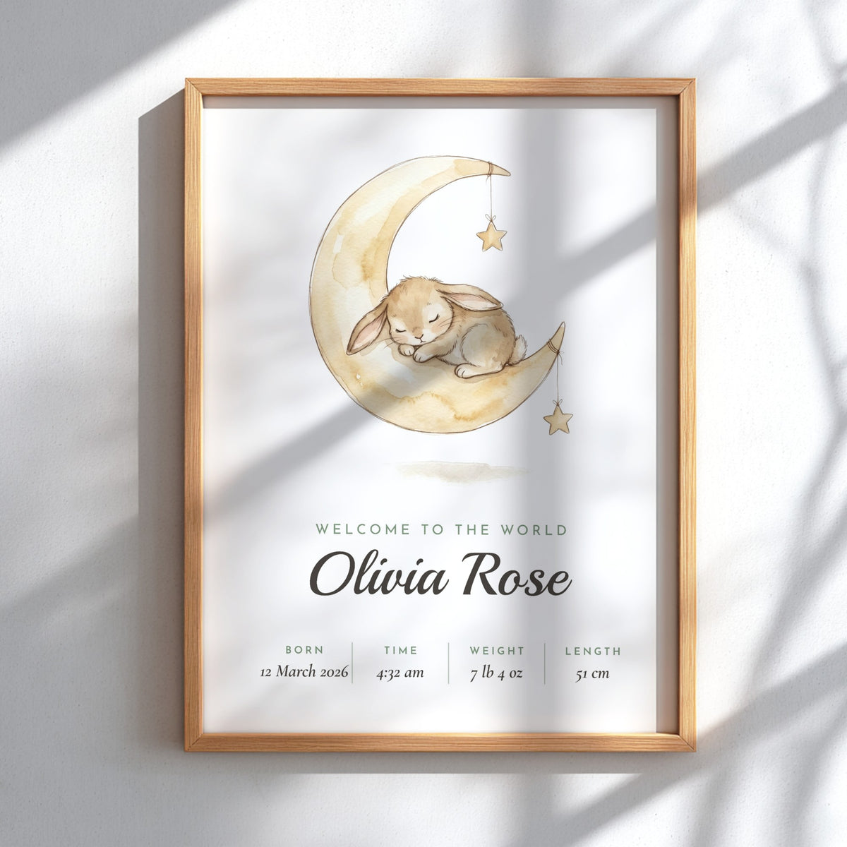 A framed personalized baby birth announcement print featuring a watercolor illustration of a sleeping bunny on a crescent moon with stars. The text reads 'WELCOME TO THE WORLD Olivia Rose' with birth details below, displayed in a light wood frame against a white wall with soft shadow overlays.