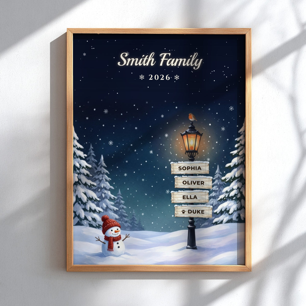 A personalized framed winter night scene print on a white wall. The artwork shows a snowy landscape under a dark blue starry sky, with a glowing streetlamp, snow-covered trees, a snowman in a red hat, and a signpost reading 'Smith Family * 2026 *' with names Sophia, Oliver, Ella, and Duke.