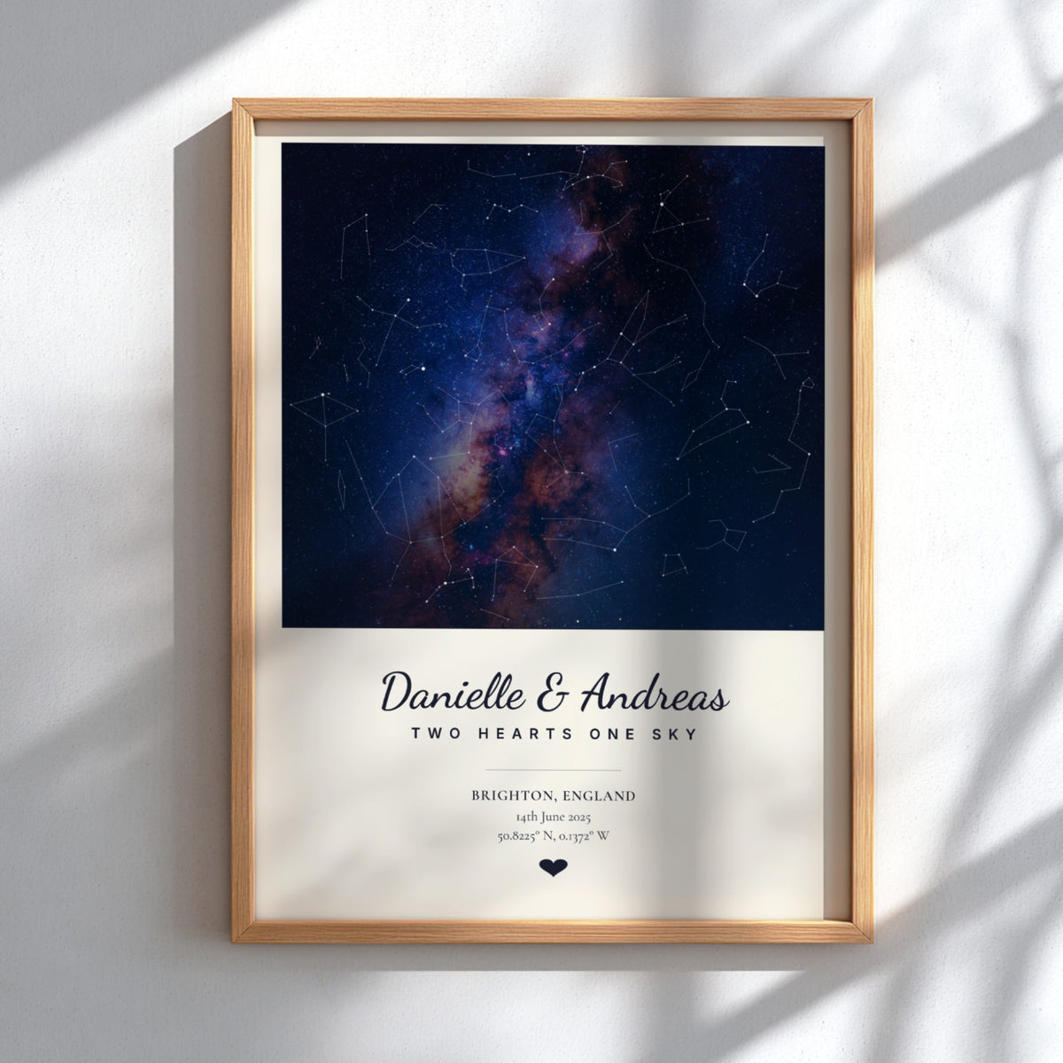 A framed personalized constellation print featuring a deep blue and purple galaxy design above a cream background with the names 'Danielle & Andreas' and wedding details, shown in a light wood frame against a white wall with soft shadow overlays.