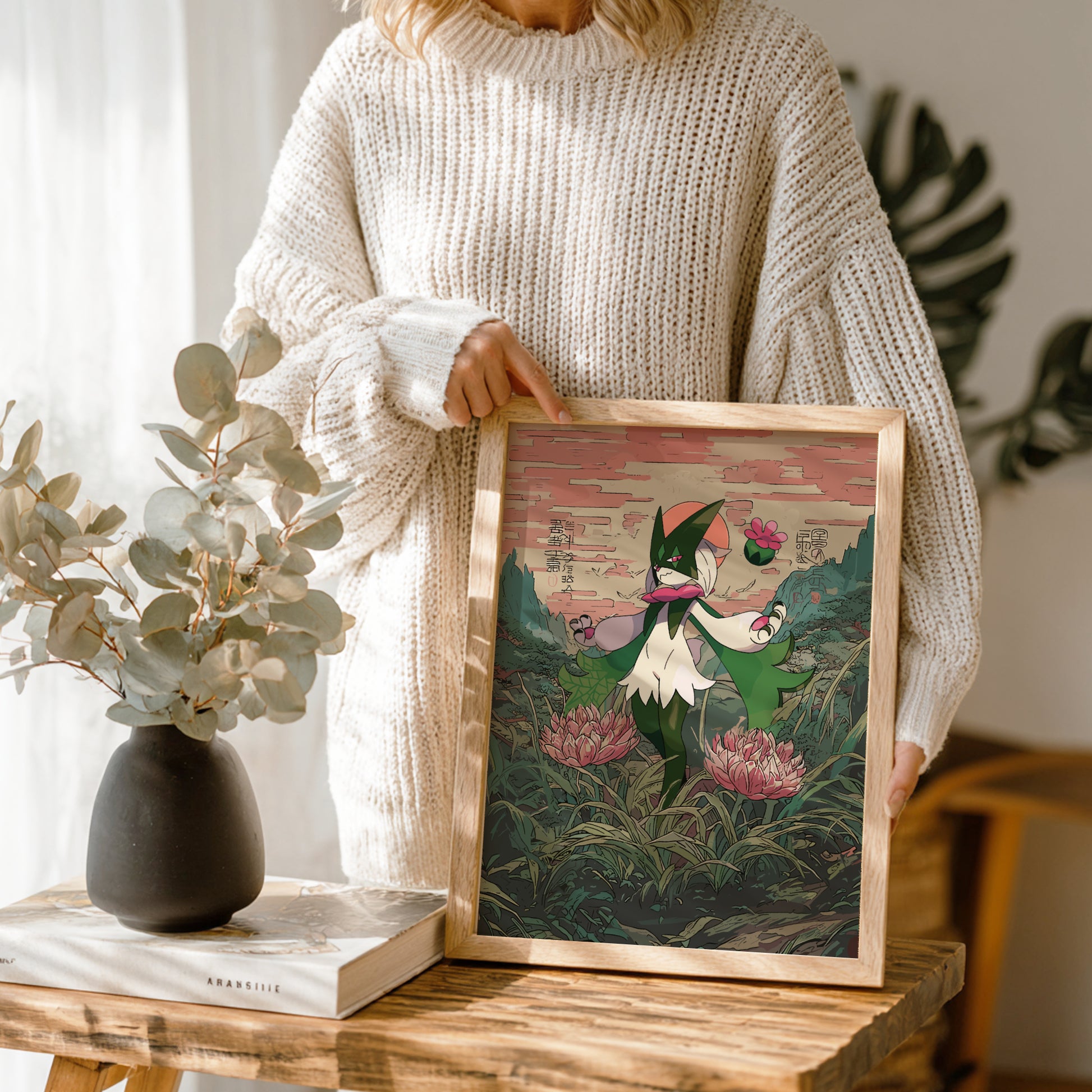 Person holding a framed artwork of Meowscarada Pokemon in a forest with a vase and book on a table.