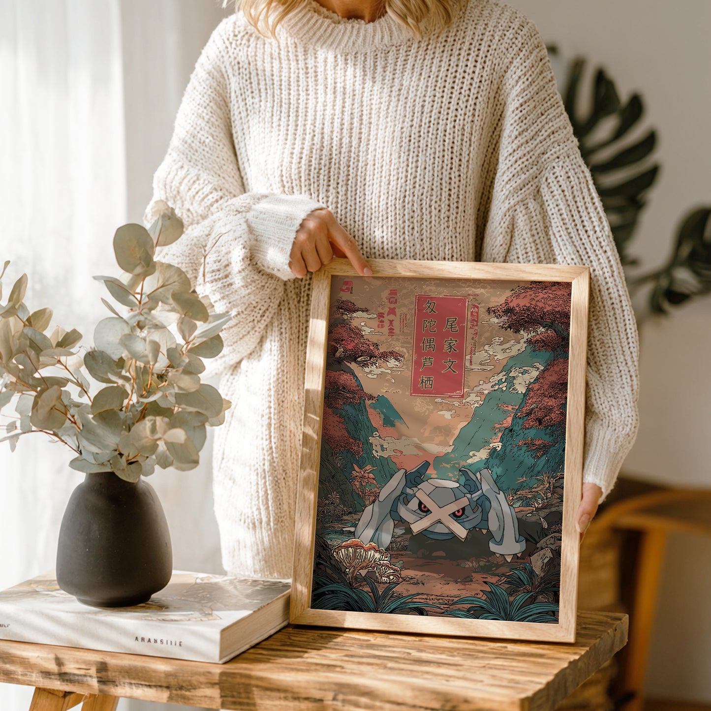 Person holding a framed artwork of Metagross Pokemon on a wooden table with a vase and book in the background.