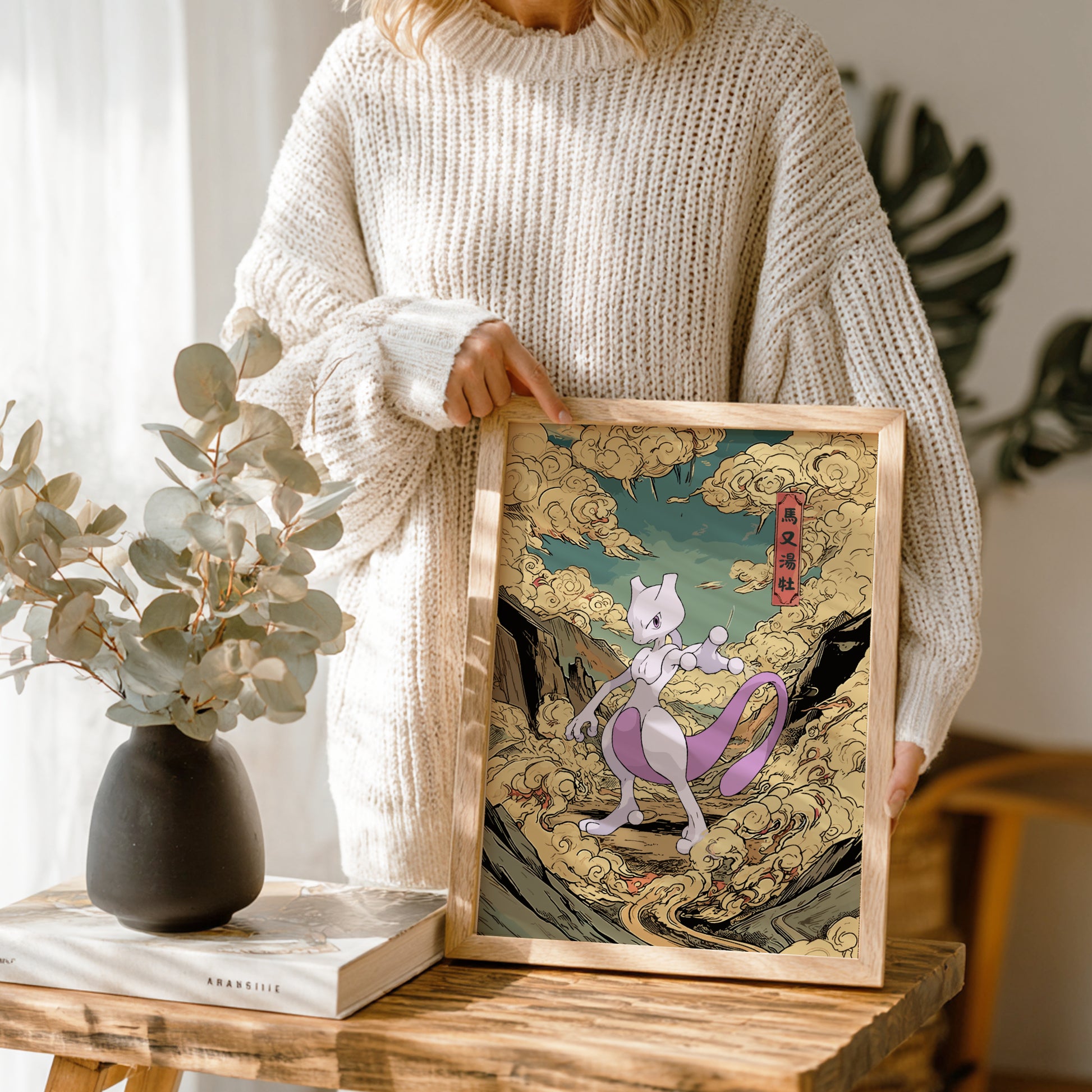 Person holding a framed artwork of Mewtwo Pokemon on a wooden table with a vase and books in the background.