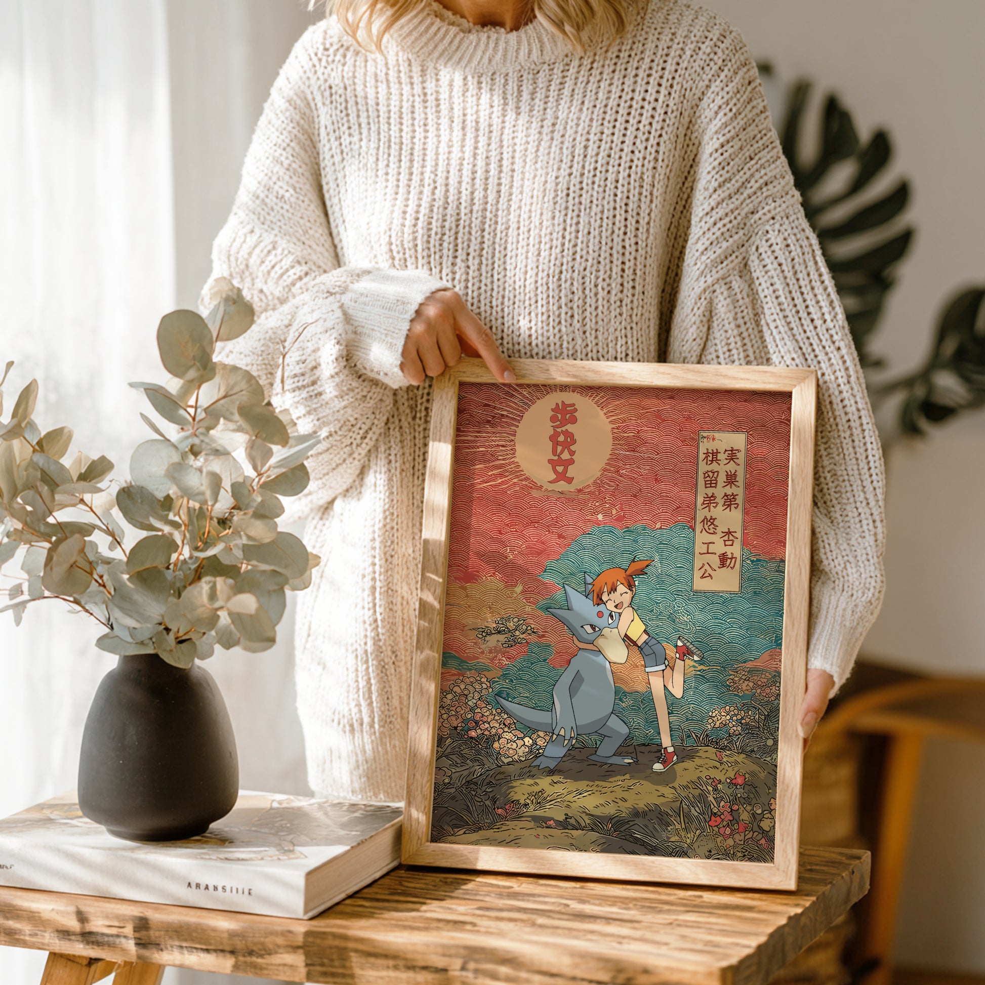 Person holding a framed artwork of Misty & Golduck Pokemon with a nature-themed design on a wooden table.