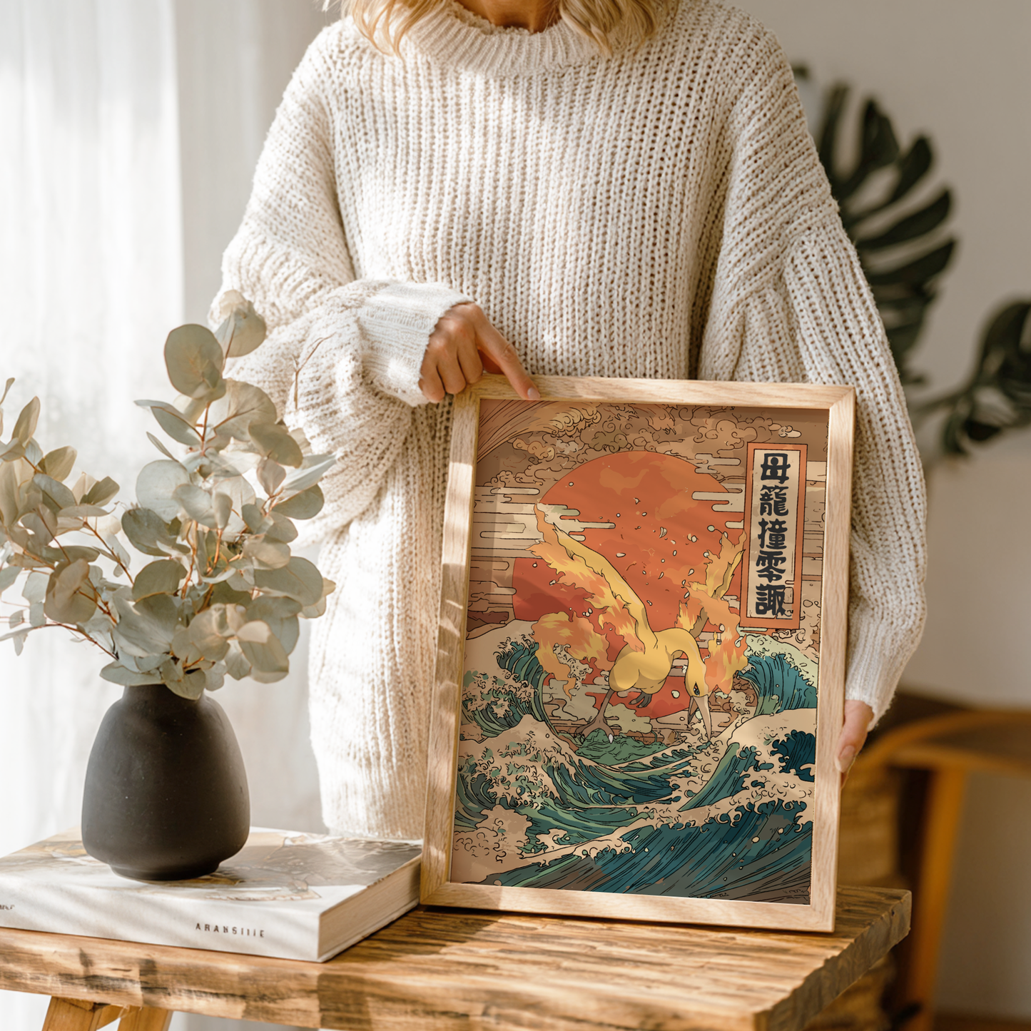 Person holding a framed artwork of Moltres Pokemon on a wave with a sunset, in a cozy indoor setting.