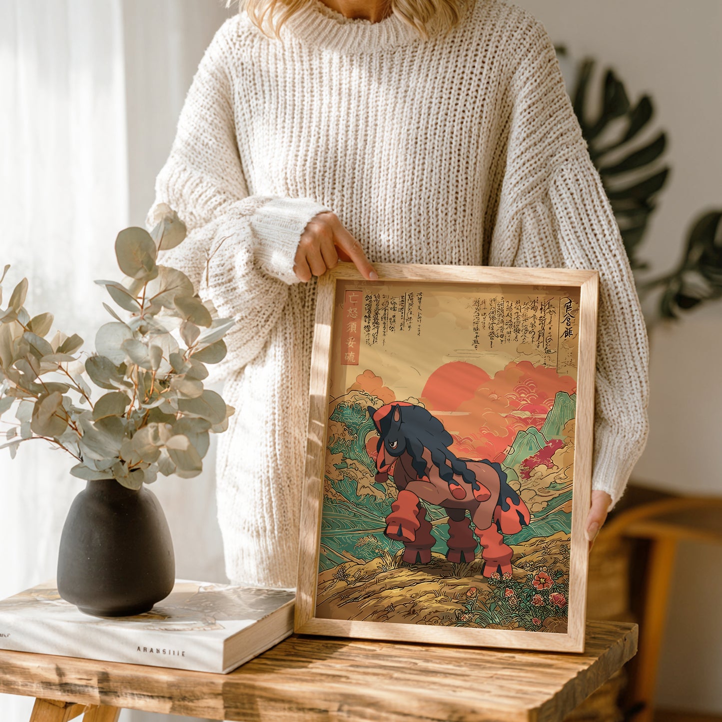 Person holding a framed artwork of a stylized Mudsdale Pokemon in a natural setting, with a vase and book on a wooden table.