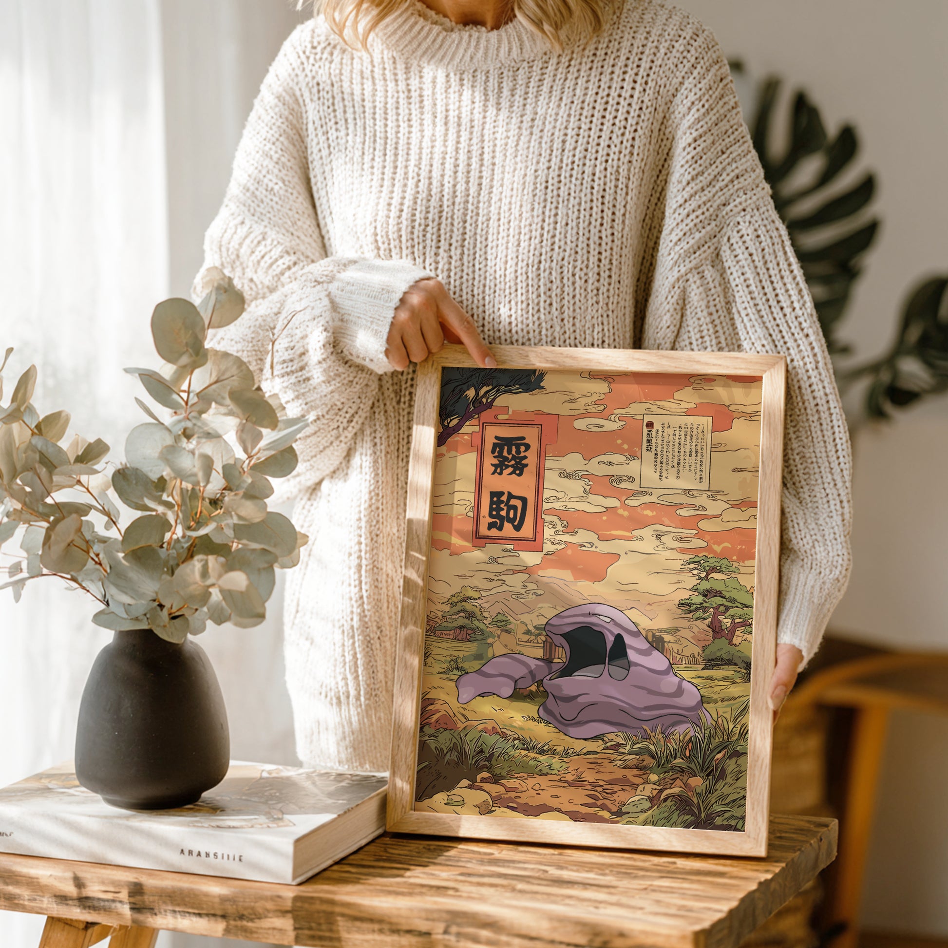 Person holding a framed artwork of Muk Pokemon on a wooden table with a vase and book in the background.