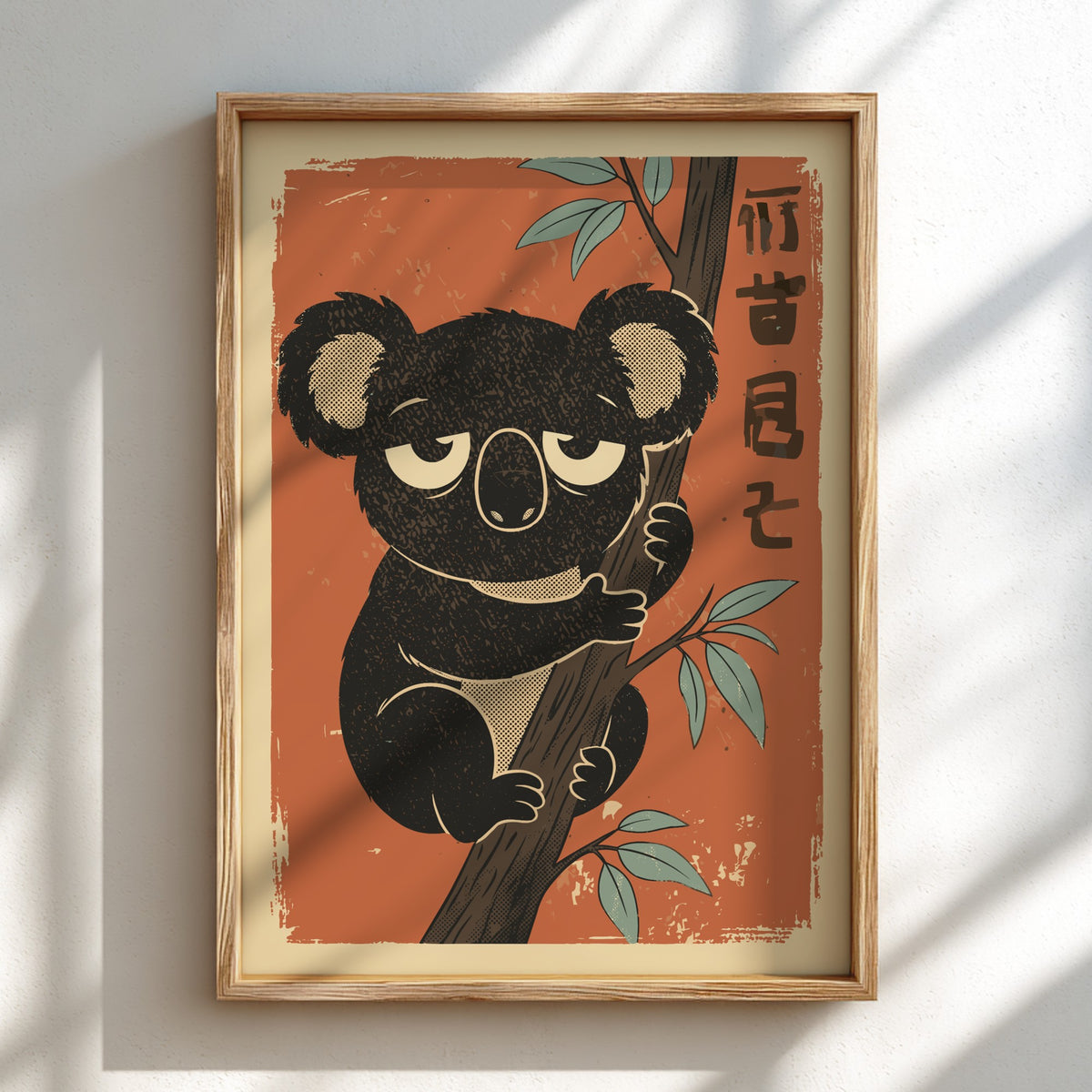 A framed art print featuring a cute, textured black koala climbing a tree against a warm orange background, mounted on a white wall with soft diagonal shadows.