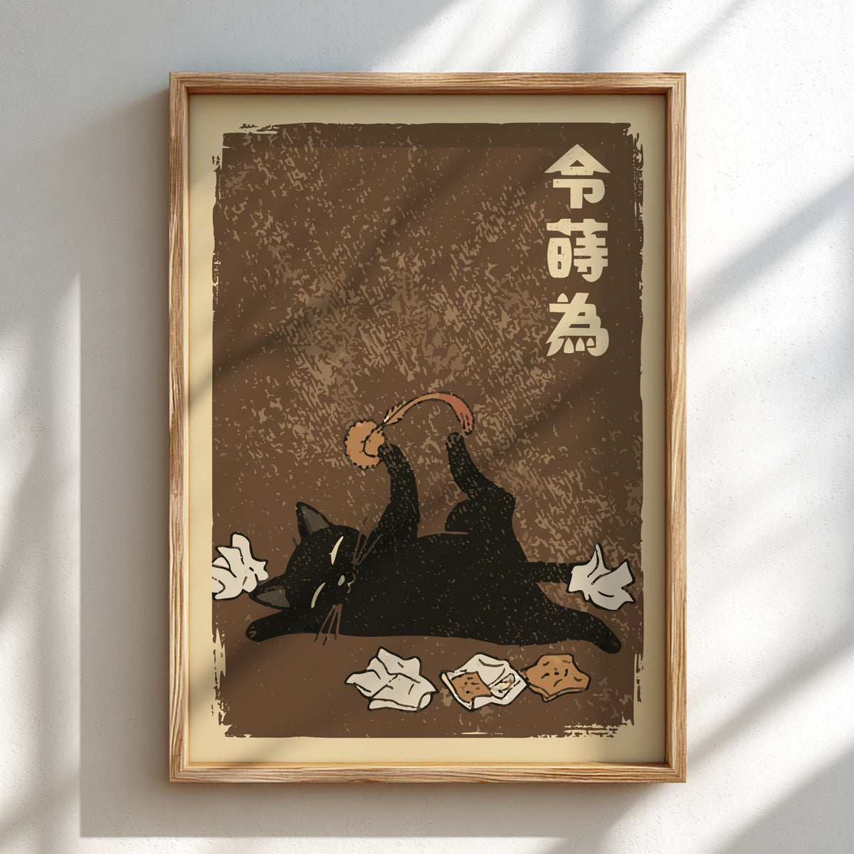 A framed art print featuring a black cat lying on its back playing with a crumb, with Japanese characters in the upper right, displayed on a light wall with soft diagonal shadows.