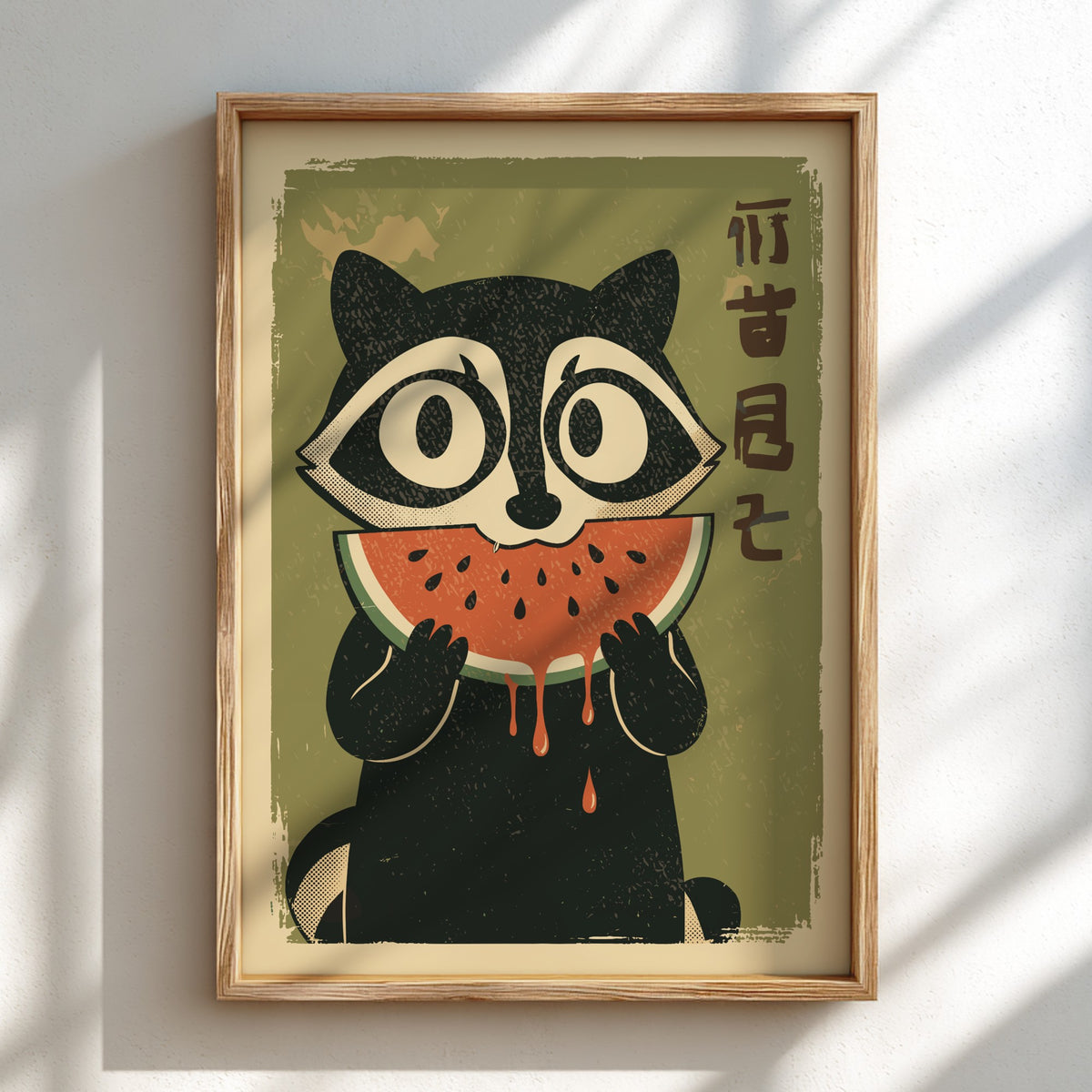 A framed art print featuring a stylized black-and-white raccoon eating a slice of watermelon, with vertical Chinese characters on the right, displayed on a light wall with soft window shadows.